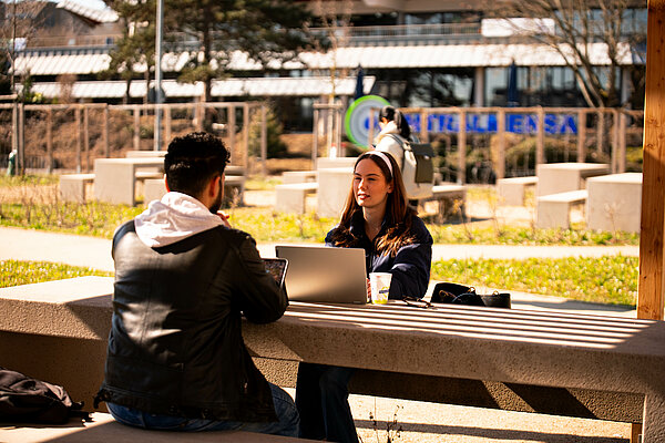 2 Persons sitting outside in front of Zentralmensa Building sign seen in the background